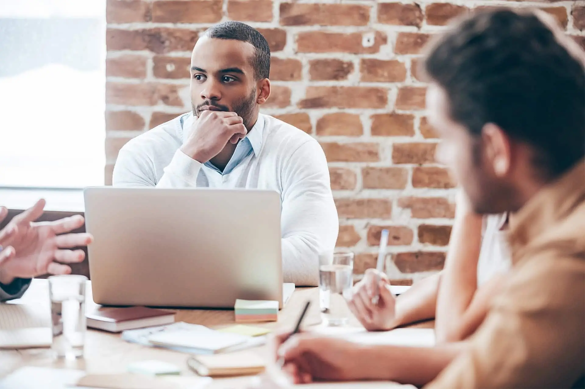 Listening to new business strategy. Thoughtful young African man holding hand on chin and listening while his coworkers discussing something sitting at the office table