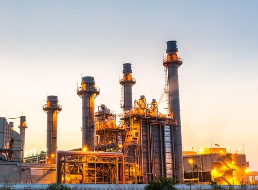 Large-scale industrial energy plant featuring tall smokestacks, complex piping, and bright yellow lights at dusk.