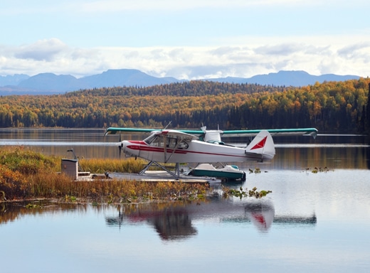A red-and-white bush plane docked on a calm Alaskan lake with autumn-colored forests and mountains